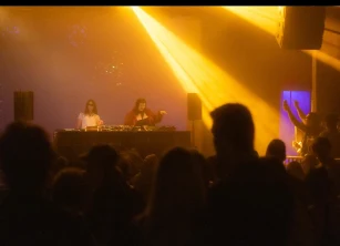 Two DJs behind the decks in amber light, crowd in silhouette